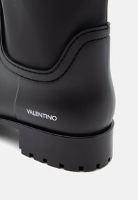 Črne gumijaste škornje s konturovano šaftom, teksturirano površino in debelim črnim podplatom. Na strani je belo "VALENTINO" blagovno oznako.