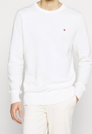 Homme portant un pull blanc texturé à col rond et manches longues avec un petit logo Tommy Hilfiger sur le côté gauche de la poitrine et un pantalon clair.