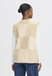 Beige vintage stila plāksteris veste, kas izgatavota no teksturētas flīsa, ar pārspīlētu griezumu un izgrieztu kakla līniju. Valkāta virs baltas krekla ar garām piedurknēm.