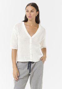 White knitted cardigan with a V-neck, short sleeves, and button front. Features a textured open weave pattern and relaxed fit.