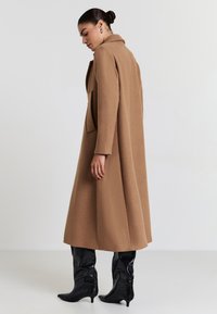 Cappotto di lana marrone con colletto a rever e tasche laterali, caratterizzato da una silhouette lunga e fluida, indossato con stivali alti neri fino al ginocchio.
