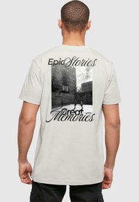 Krém színű pamut póló, fekete-fehér grafikai nyomattal, amely városi tájat és a "Epic Stories" és "great Memories" feliratokat ábrázol.