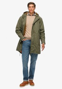Parka verde oliva con capucha, forro interior texturizado, combinada con suéter beige, camisa de cuadros, jeans azules y zapatos marrones con cordones.