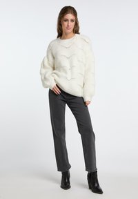 Maglione bianco lavorato a maglia grosso con un motivo ondulato, maniche lunghe, abbinato a jeans grigi a gamba dritta e stivali neri alla caviglia.