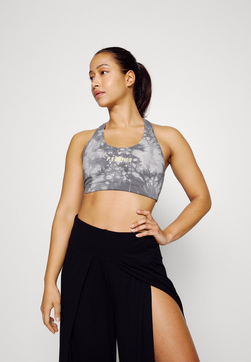 Sutiã desportivo cinza com padrão tie-dye e logo P.E Nation, combinado com calças pretas de cintura alta com um corte lateral, tecido suave e um ajuste relaxado.