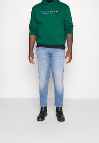 Grøn hættetrøje med hvidt logo, båret over en sort skjorte. Lyseblå jeans og sorte ankelstøvler fuldender outfittet.
