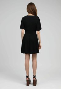 Robe noire courte avec une taille cintrée, une jupe évasée et des manches courtes ; fabriquée en tissu texturé ; portée avec des sandales à talons épais.