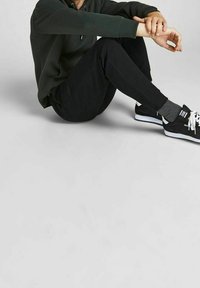 Groene hoodie met trekkoord, zwarte trainingsbroek, grijze sokken en zwarte sneakers met witte accenten, zittend op een lichtgrijze achtergrond.