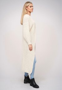 Jasno beżowy sweter z dzianiny z długimi rękawami, otwartym przodem i bocznymi rozcięciami; zestawiony z jasnoniebieskimi dżinsami i czarnymi botkami za kostkę.