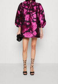 Kvinna klädd i en kort svart klänning med stora rosa blommönster, puffärmar, som håller en svart clutch och bär svarta sandaletter med remmar.