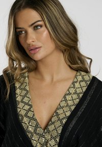 Prenda negra con un escote en V profundo, que presenta un patrón geométrico dorado y acentos metálicos a lo largo del escote. Textura suave.