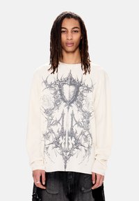 Νεαρό άτομο με μακριές πλεξούδες φοράει κρεμ μακρυμάνικο πουκάμισο με περίπλοκο σκούρο αφηρημένο σχέδιο στο μπροστινό μέρος και μαύρο παντελόνι.