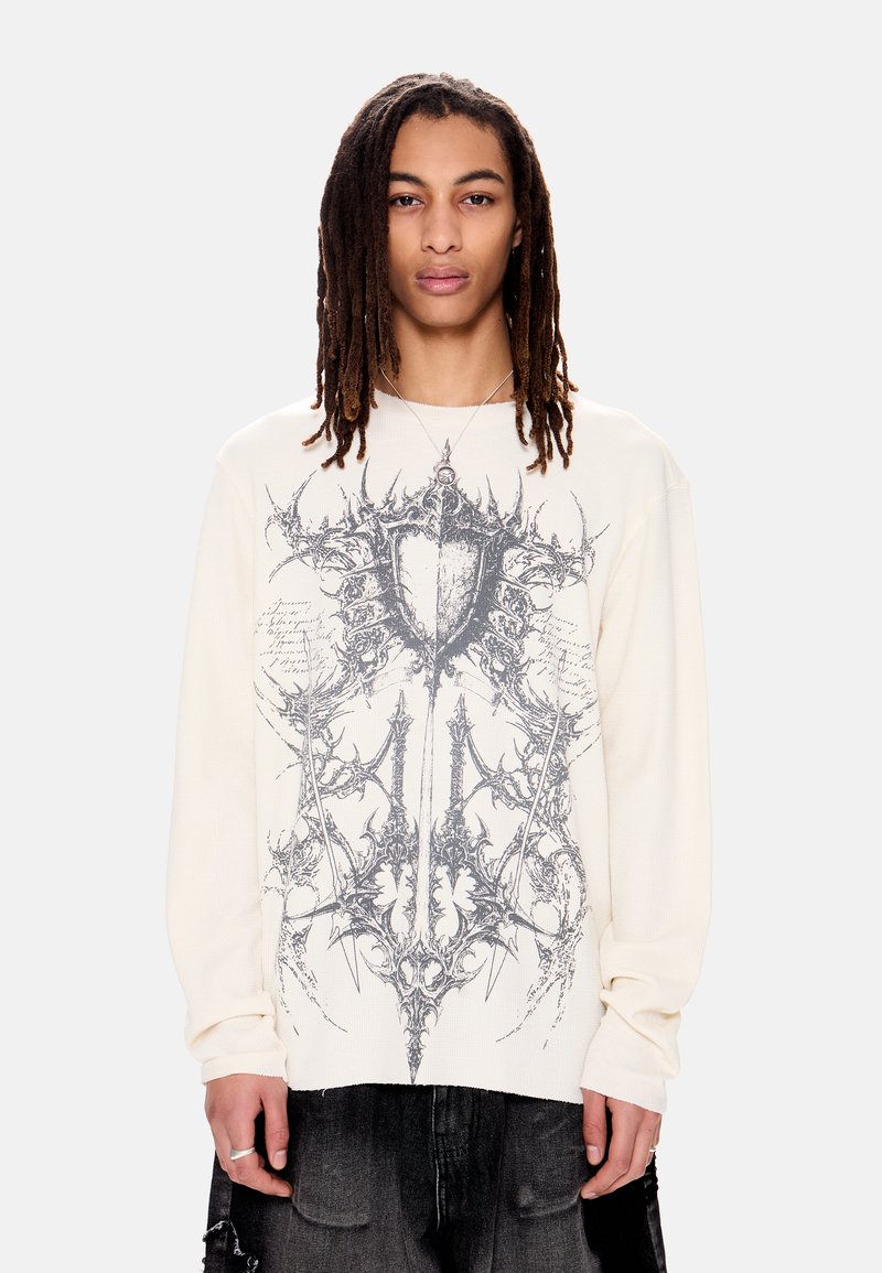 Νεαρό άτομο με μακριές πλεξούδες φοράει κρεμ μακρυμάνικο πουκάμισο με περίπλοκο σκούρο αφηρημένο σχέδιο στο μπροστινό μέρος και μαύρο παντελόνι.