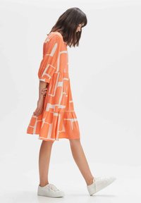 Oranges Kleid mit weißen geometrischen Mustern, Dreiviertelärmel, einem schichtweisen Rock und einer lockeren Passform, kombiniert mit weißen Sneakern.
