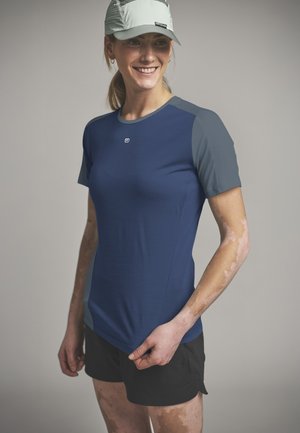 Frau mit grauer Mütze, dunkelblauem und grauem Sportshirt sowie schwarzen Shorts, die lächelt und seitlich schaut vor neutralem Hintergrund.