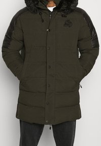 Chaqueta acolchada verde oliva con capucha de piel sintética desmontable, detalles en cuero negro en los hombros y cierre frontal con botones; textura acolchada.