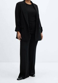 Sort skræddersyet blazer over en tætsiddende top og viddebukser, alle i ensartet sort stof. Har rene linjer og minimalistisk design.