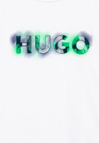 Bílé tričko s nápisem "HUGO" tučným písmem, s přechodem zelené a šedé barvy a mírně texturovaným povrchem.