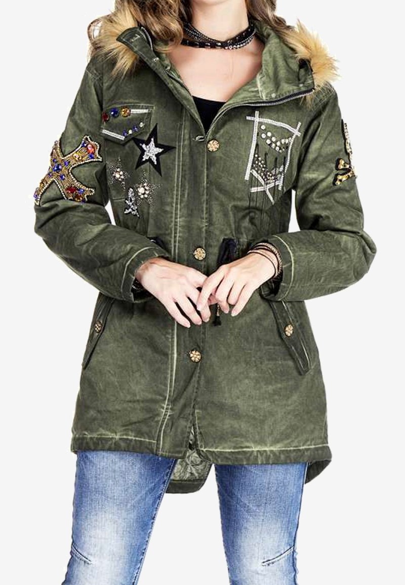 Femme portant une parka verte avec capuche bordée de fourrure, ornée de patchs brodés en forme d'étoile et de croix ainsi que de perles, associée à un jean bleu.