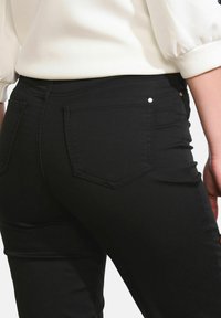 Donna che indossa un paio di jeans neri attillati che mostrano le tasche posteriori e una blouse bianca infilata con maniche a palloncino.