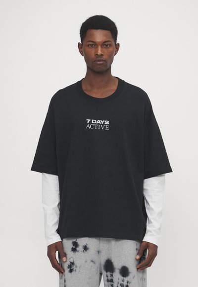 T-shirt en coton noir, coupe ample, manches courtes, avec "7 DAYS ACTIVE" imprimé en blanc. Porté par-dessus une chemise blanche à manches longues.