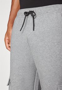 Zbliżenie osoby noszącej jasnoszare spodnie dresowe z czarnym sznurkiem wiązanym na kokardę w talii, ręka spoczywa obok biodra.