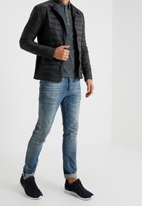 Zwarte, gequiltte jas over een donkergrijze polo, gecombineerd met lichtblauwe versleten jeans en donkere sneakers met witte zolen. Casual outfit.
