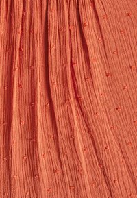 Textilní látka v texturované, rezavě oranžové barvě s vystouplými vzory teček a sklady, vytvářející měkký, vlnící se vzhled.