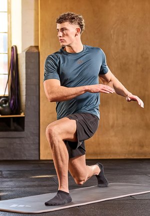 Athletic male model in a teal short-sleeve shirt and black shorts performing a lunge on a gray exercise mat. Textured fabric, streamlined design.