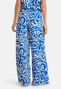 Weite Hosen aus cremefarbenem Stoff mit einem blauen abstrakten Muster. Das Design umfasst verschiedene organische Formen und sanfte Texturen.