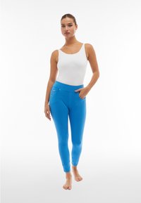 Leggings blu con vita alta, realizzati in tessuto elastico, con tasche laterali e texture liscia. Abbinati a un top bianco senza maniche.
