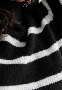 Personne portant un pull en tricot à rayures noires et blanches, avec des cheveux bouclés foncés en partie visibles dans la partie supérieure du cadre.