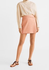 Beige pitkähihaisen paidan, jossa on brodeeratut yksityiskohdat, yhdistäminen vaaleanpunaiseen mini-hameeseen, jossa on kolme nappia. Yhdistetty ruskeisiin tasapohjaisiin sandaaleihin.