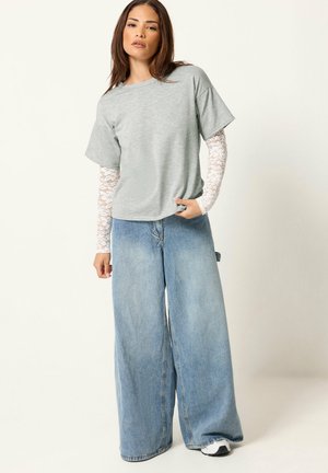Kvinde iført lysegrå T-shirt over hvide blondeærmer, blå vidde bukser og hvide sneakers, stående mod en ensfarvet baggrund.