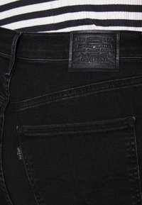 Jeans de mezclilla negra con un parche de cuero con texto en relieve y detalles de costura. El diseño incluye bolsillos traseros estándar y una etiqueta.