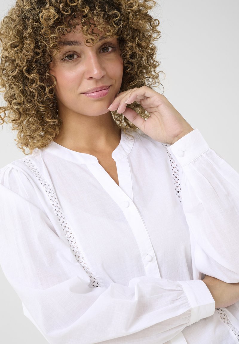 Camicia bianca con bottoni, caratterizzata da maniche a sbuffo, dettagli in pizzo sulle spalle e colletto alla cinese. Tessuto morbido e leggero.