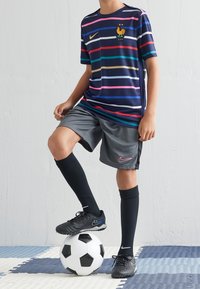 Niño con camiseta de fútbol de Francia a rayas y pantalones cortos grises, de pie con un pie sobre un balón de fútbol negro y blanco.