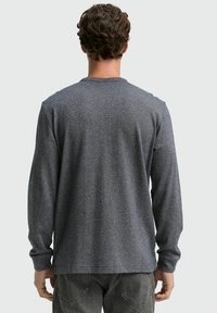 Graues Langarmshirt aus strukturiertem Stoff, mit Rundhalsausschnitt und Seitenschlitzen. Die Rückseite hat einen klassischen Schnitt.