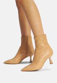 Stivaletti beige realizzati in pelle liscia con punta affusolata, cerniera laterale e tacco triangolare. Presentano un polsino aderente e cuciture sottili.