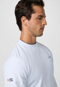 Camisa blanca de manga larga con cuello alto, que presenta un pequeño logo rojo y negro en el pecho y en la manga izquierda. Textura suave.