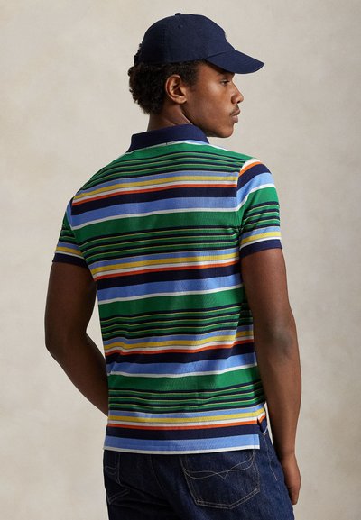 Ung mann iført en flerfarget stripete polo-skjorte, marineblå caps og mørk jeans, vendt bort med hodet vridd til siden.