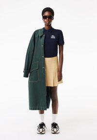 Abrigo verde con costuras visibles, camiseta polo azul marino con logo, falda de tablas beige, calcetas blancas, zapatillas negras y beige con estampado, y gafas de sol.