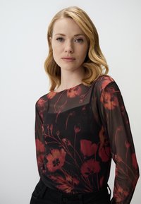 Blouse noire transparente avec un motif floral rouge, manches longues, encolure arrondie et texture lisse. Associée à un pantalon sombre.