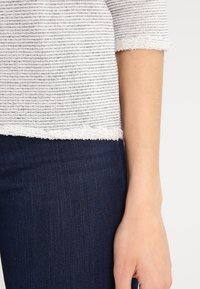 Maglione a righe grigie e bianche con orlo grezzo, abbinato a jeans di denim blu scuro. Primo piano di manica e polso visibili.