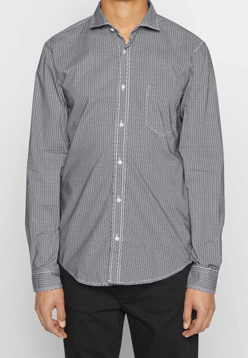 Homme portant une chemise à manches longues boutonnée avec un petit motif géométrique noir et blanc et un pantalon noir, debout devant un fond uni.