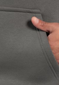 Tejido de sudadera gris con detalles de bolsillo, presenta un punto texturizado y costuras visibles; una mano está colocada en el bolsillo.