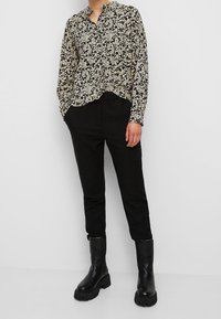 Personne portant un chemisier à manches longues noir à motifs floraux, un pantalon noir avec une poche latérale, et des bottes noires épaisses, debout devant un fond uni.