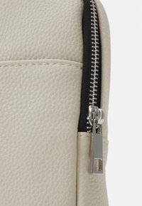 Beige textured backpack material with a black zipper detail. Features a horizontal pocket and metal zipper pull accent.