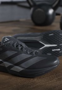 Μαύρα και γκρίζα παπούτσια τρεξίματος Adidas με σόλα Lightstrike Pro σε ξύλινο πάτωμα, ασαφή kettlebells στο φόντο.