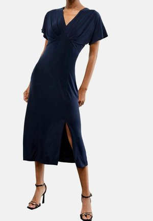 Femme portant une robe midi bleu marine à manches papillon, encolure en V, fente sur le devant et sandales noires à talons hauts.